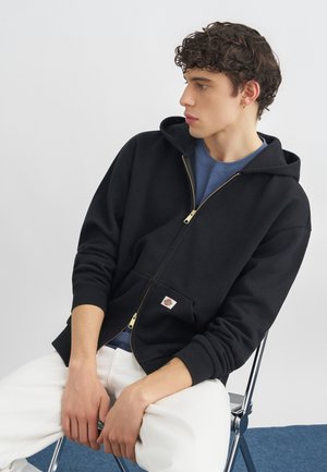 Ung mann med krøllete hår iført en svart hettejakke med glidelås, blå skjorte, hvite bukser og gull hoop-øredobb, sittende på en metallstol.