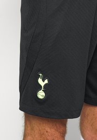 Svarta sportshorts med en strukturerad yta och en framträdande grön logotyp på vänstra benet, som föreställer en fågel ovanför en boll.