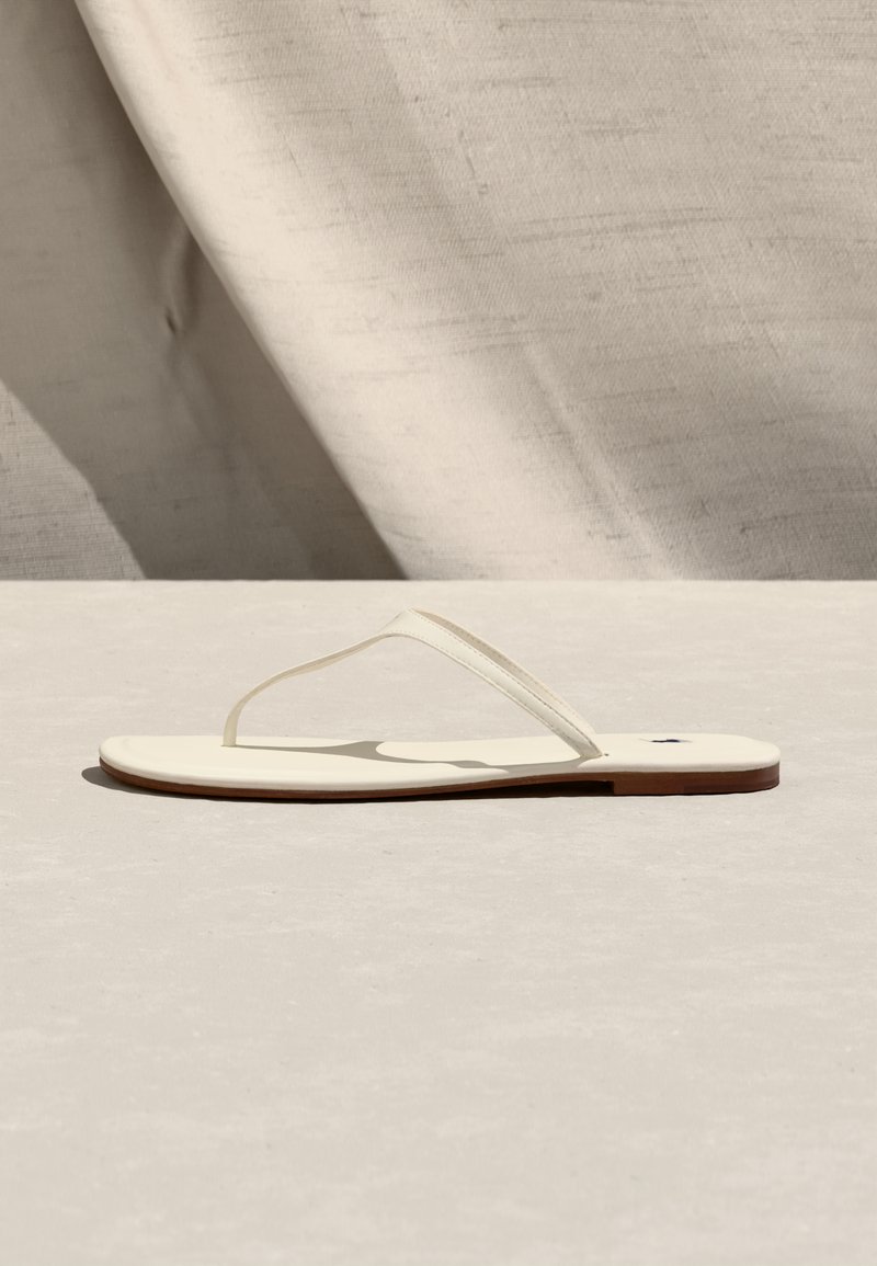 Μινιμαλιστικό σανδαλάκι κρεμ με λεπτούς ιμάντες και επίπεδη σόλα, τοποθετημένο σε φωτεινή υφή επιφάνεια με φόντο από ριγμένο ύφασμα.