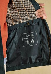 Interiorul unei jachete verde închis, având o căptușeală în carouri bej și bleumarin. Include o etichetă neagră cu logo-ul "JOULES" și simboluri pentru impermeabilitate.