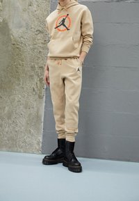 Beige Kapuzenpullover mit schwarzem Logo und orangefarbenem Kreis, kombiniert mit beige Jogginghosen und schwarzen Schnürstiefeln. Glattes Material, lässige Passform.