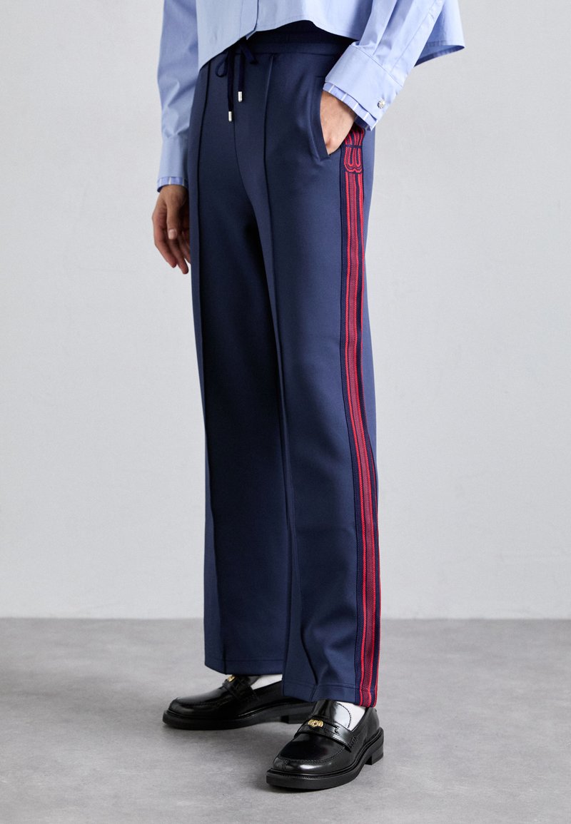 Pantalones de chándal navy con franjas rojas a los lados, cinturilla elástica y bolsillos laterales. Combinados con zapatos negros y una camisa de botones azul claro.