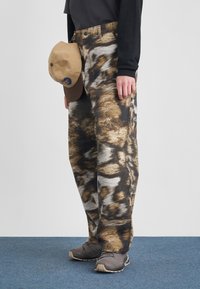 Άτομο με παντελόνι σε καφέ και μαύρο σχέδιο, γκρι αθλητικά παπούτσια, κρατώντας ένα μπεζ και καφέ καπέλο μπέιζμπολ με μαύρη μπλούζα μακρύ μανίκι.