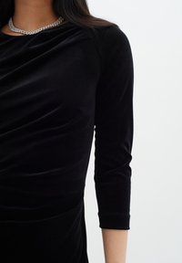 Černý sametový top s třičtvrtinovými rukávy, s volně splývající látkou a stříbrným ozdobným náhrdelníkem, který zvýrazňuje výstřih.