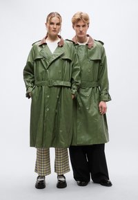 Πράσινα δερμάτινα παλτό τύπου τρεντς με καφέ γιακάδες, διπλή σειρά κουμπιών και ζώνη στη μέση, φορεμένα πάνω από παντελόνια με σχέδια και μονόχρωμα.