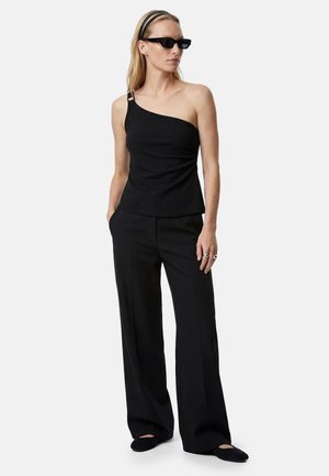 Femme portant un top noir à une épaule, un pantalon large noir, des lunettes de soleil noires, des ballerines noires et un bandeau noir sur fond uni.