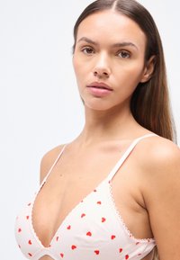 Τριγωνικό σουτιέν από απαλό ύφασμα με ροζ βάση, διακοσμημένο με μικρές κόκκινες καρδιές και κυματιστές άκρες, με λεπτές ρυθμιζόμενες τιράντες.