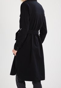 Osoba ubrana w długi czarny płaszcz z paskiem, z zapiętymi rękawami, stojąca z plecami do widza, na jednolitym jasnym tle.