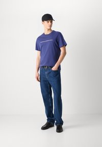 Purple cotton t-shirt with "BARBOUR INTERNATIONAL" text, paired with light blue denim jeans and black shoes. Black cap completes the look.