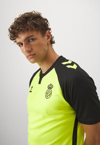 Maglietta sportiva di un brillante giallo neon con dettagli neri. Presenta maniche corte, scollatura a V e un logo stampato sul davanti. Tessuto testurizzato.