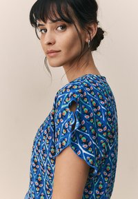 Blouse bleue à motifs avec des designs floraux en orange et vert. Manches courtes avec détails découpés. Texture douce et coupe décontractée.