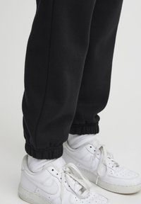 Crne trenirke s elastičnim manžetama u kombinaciji s bijelim Nike Air Force 1 tenisicama, koje imaju glatku kožnu teksturu i perforirane detalje.