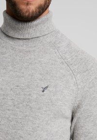 Homme portant un pull à col roulé côtelé gris clair avec un petit logo d'oiseau brodé foncé sur la poitrine gauche.