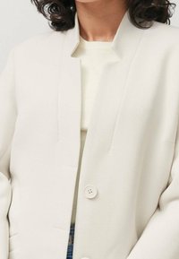 Chaqueta color crema con un diseño estructurado, cuello de muesca y dos botones, llevada sobre un suéter ligero. Tejido texturizado con acentos mínimos.