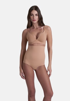 Vrouw in een huidskleurige beha en high-waisted shapewear slips, staand tegen een effen lichte achtergrond.
