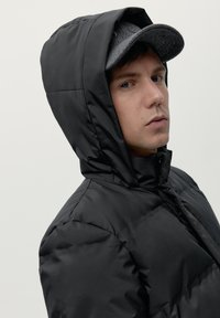 Melnā pūkaina jaka ar kapuci, ar gludu, spīdīgu apdari. Modelis zem kapuces valkā pelēku filca cepuri.