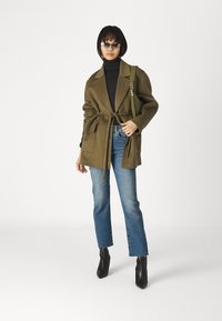 Abrigo verde oliva con cinturón y grandes bolsillos, combinado con un cuello de tortuga negro, jeans azules y botas negras hasta el tobillo. Los accesorios incluyen gafas de sol y un bolso verde.