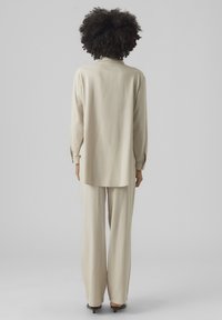 Beiges Zweiteiler-Outfit mit einem langärmeligen Oberteil mit abgerundetem Saum und lockeren Hosen, beide aus einem glatten, leichten Stoff.