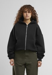 Schwarze Zip-Hoodie mit großer Kapuze, Fronttaschen und lockerer Passform; aus weichem Material mit Rippbündchen und Saum. Kombiniert mit olivgrünen Hosen.