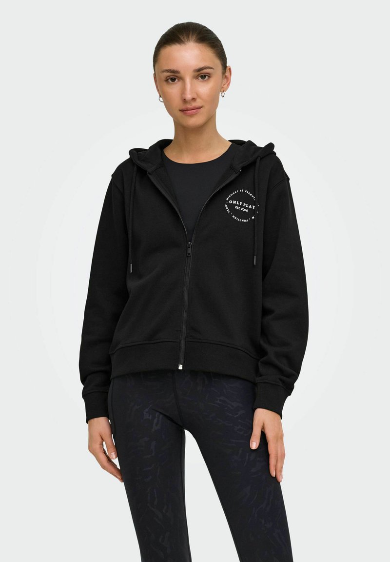 Jeune femme aux cheveux foncés portant un sweat à capuche noir zippé avec logo et des leggings noirs à motifs, debout devant un fond uni.