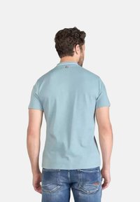 Lichtblauwe poloshirt met een klassieke kraag, korte mouwen en een klein accent van de Franse vlag op de achterkant van de nek. Draagt bij blauwe spijkerbroeken.