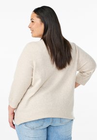 Beige gestrickter Pullover mit Dreiviertelärmeln, weichem Griff und abgerundetem Saum, kombiniert mit hellblauen Jeans.