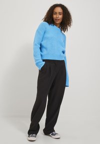 Svetlo modra pletena majica z V-izrezom in dolgimi rokavi, v kombinaciji z visokimi črnimi hlačami in modrimi športnimi čevlji.