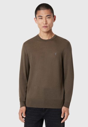 Mand med kort hår iført en brun langærmet sweater med lille logo på brystet og sorte bukser, stående mod en ensfarvet baggrund.