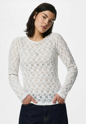 Jeune femme aux cheveux foncés portant un haut blanc en dentelle florale à manches longues et un pantalon foncé, debout les mains dans les poches.