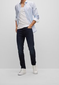 Camisa de botón azul claro, camiseta blanca, jeans oscuros, zapatillas blancas, estilo casual, con mangas remangadas y una silueta ajustada.