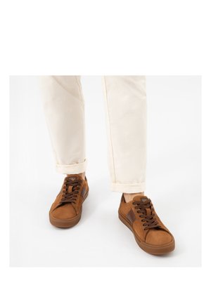 Persona che indossa pantaloni beige e sneakers basse marroni con lacci marrone scuro, in piedi contro uno sfondo bianco.