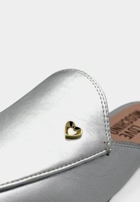 Ασημένιο δερμάτινο παπούτσι με εμφανείς ραφές και ένα μικρό χρυσό κενό καρδιάς διακοσμητικό κοντά στην μύτη.