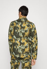 Kampanijacka i grön, gul och ljusblått tyg. Har en klassisk krage, långa ärmar och en avslappnad passform med detaljer på bakfickan.