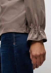 Šedý, měkký látkový top s nařasenými rukávy a hladkou texturou, v kombinaci s tmavě modrými džíny s viditelným šitím na kapsách.