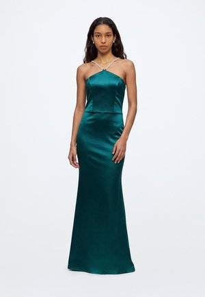 Femme debout portant une longue robe en satin bleu sarcelle foncé près du corps avec des fines bretelles croisées et un décolleté droit sur un fond blanc uni.
