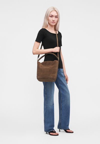 Jeune femme aux longs cheveux blonds portant un haut noir, un jean bleu à jambes larges, des sandales noires à talons, portant un grand sac d'épaule en daim marron.