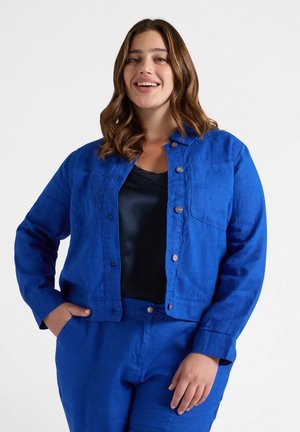 Blaue Leinenjacke mit Brusttaschen, Knopfverschluss und langen Ärmeln, kombiniert mit einem schwarzen Oberteil. Glatte Textur und leuchtende Farbe.