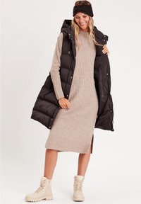 Gilet noir en duvet sur une robe en tricot côtelé beige avec des fentes sur les côtés, associé à des bottes beiges à lacets. Accessoire bandeau noir inclus.