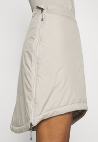 Jupe beige matelassée avec fermeture éclair à l'avant, texture lisse et forme légèrement évasée, avec un ourlet arrondi.