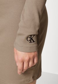 Kjol med lång ärm i ribbat tyg i beige med svart broderad "cK"-logotyp på ärmsluten. Har en figurnära design och slät textur.