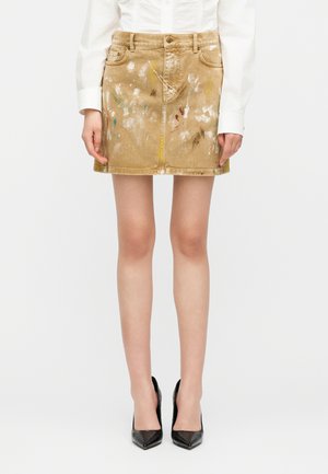 Beige mini skirt with paint splatters worn with white blouse and black high-heeled shoes, shown from waist to feet.