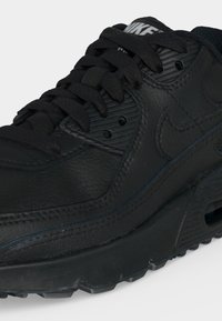 Čierny teniska Nike Air Max s textúrovanou kožou, hrubou podrážkou, Nike logom na boku a čiernymi šnúrkami na jednofarebnom pozadí.
