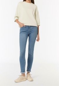 Vrouw met een beige lange mouwen top, lichtblauwe skinny jeans met omgeslagen pijpen en beige chunky sneakers, staand tegen een witte achtergrond.