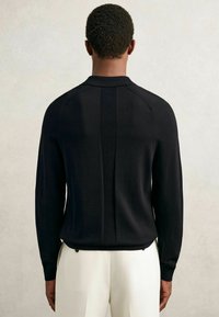 O persoană care poartă un pulover negru cu mânecă lungă, cu dungi verticale nervurate pe spate, asortat cu pantaloni albi, întors cu spatele.