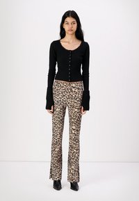 Svart ribbet langermet topp med knappelukking; kombinert med beige leopardmønstrede bukser med vide ben. Modellen står med hendene langs siden.