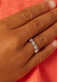 Zilveren ring met bloemmotief ligt op een hand met een bruine huid, tegen een textuur achtergrond in koraalkleur. De ring heeft kleine, ronde bloemaccenten.