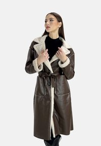 Brun læderjakke med krave i cremefarvet faux fur, bælte i taljen og to forreste lommer. Glat tekstur med kontrasterende syninger.