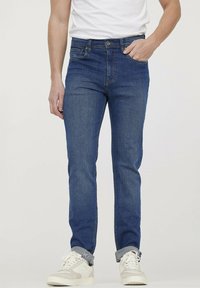 Blauwe spijkerbroek met een slim fit, voorzien van vijf zakken, een ritsgulp en een lichte vervaging; gedragen met witte en grijze sneakers.