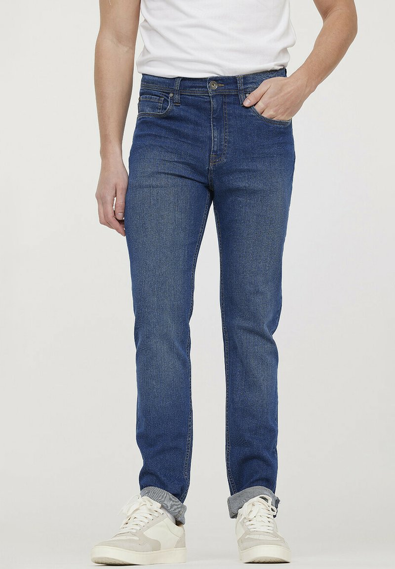 Blauwe spijkerbroek met een slim fit, voorzien van vijf zakken, een ritsgulp en een lichte vervaging; gedragen met witte en grijze sneakers.