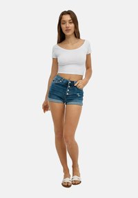 Camiseta blanca corta, pantalones cortos de mezclilla azul con cintura alta y puños enrollados, cierre frontal de cinco botones y detalles de bolsillo. Sandalias blancas de tipo slide.